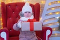 Portrait little girl whos sits in a red christmas chair and gives a Christmas gift outside.