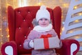 Portrait little girl whos sits in a red christmas chair and holds Christmas gift outside.