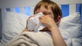 Portrait of little sick boy with runny nose lying in bed and blowing nose in paper tissue. Concept of children illness