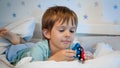 Portrait of little smiling toddler boy lying in bed at night and playing with two wooden toy cars before going to sleep. Royalty Free Stock Photo