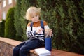 Portrait of little student with backpack, notepad and bottle of water sitting near school building. Child drink water. Kids back