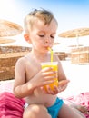 Portrait of little toddler boy sitting on hte sun bed at sea beach and drinking orange juice from straw Royalty Free Stock Photo