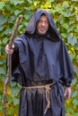 Portrait of a lone monk with a wooden staff against a neutral background of grape leaves.