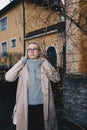 Portrait of long haired blonde woman wearing a sand coat with wool sweater, and black jeans, standing behind old fashioned house. Royalty Free Stock Photo