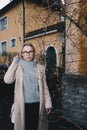 Portrait of long haired blonde woman wearing a sand coat with wool sweater, and black jeans, standing behind old fashioned house. Royalty Free Stock Photo