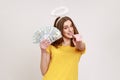 Portrait of lottery winner lucky angelic girl with halo over head showing fan of money, holding dollar bills and pointing to Royalty Free Stock Photo