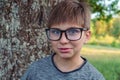 Loveable boy with blue eyes, in glasses in gray longsleeve standing in front of tree in park and enjoying fresh air. Royalty Free Stock Photo