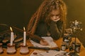 Little witch dressed in dark clothes sitting at the table against a black smoky background. There are magic wand, books Royalty Free Stock Photo