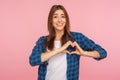 Portrait of lovely romantic girl in checkered shirt showing heart shape with hands and smiling at camera Royalty Free Stock Photo