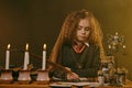 Little witch dressed in dark clothes sitting at the table against a black smoky background. There are magic wand, books Royalty Free Stock Photo