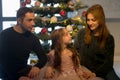 Parents hug their adorable daughter, mom and dad, sitting near the Christmas tree. Royalty Free Stock Photo