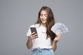 Portrait of lucky young woman smiling while holding fan of money and cell phone in hands over gray background