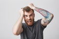 Portrait of mad beautiful bearded man with tattoed arm and stylish hairstyle in casual gray shirt tears hair with hands