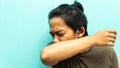 A portrait of a Malay man coughing and sneezing to his elbow neck with closing both eyes on isolated blue background