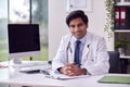 Portrait Of Male Doctor Or GP Wearing White Coat At Desk In Office With Clipboard Royalty Free Stock Photo