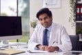 Portrait Of Male Doctor Or GP Wearing White Coat At Desk In Office Writing Notes On Clipboard Royalty Free Stock Photo