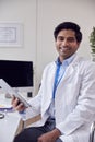 Portrait Of Male Doctor Or GP Wearing White Coat Sitting At Desk In Office Using Digital Tablet Royalty Free Stock Photo