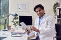 Portrait Of Male Doctor Or GP Wearing White Coat Sitting At Desk In Office Using Digital Tablet Royalty Free Stock Photo