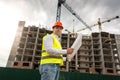 Portrait of male engineer in hardhat and safety with blueprints standing against working building cranes Royalty Free Stock Photo