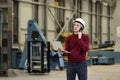 Portrait of a male factory manager in a white hard hat and red sweater holding laptop and mobile phone.
