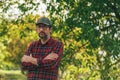 Portrait of male farmer with arms crossed wearing plaid shirt and trucker`s hat posing in walnut orchard and looking at camera