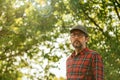 Portrait of male farmer wearing plaid shirt and trucker`s hat posing in walnut orchard and looking at camera