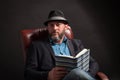 Portrait of man with beard and hat sitting on leather chair reading book. Royalty Free Stock Photo