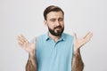 Portrait of a man with a beard raising his hands looking uncertain, standing near white wall. Husband tries to explain