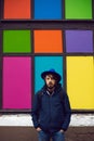 portrait of a man with a beard standing in a jacket and blue hat stands against a background of colorful rectangle