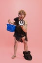 Portrait of man in character of neanderthal, primitive person shouting in megaphone and holding shopping basket isolated