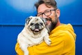 Portrait of man and dog. Young adult handsome with beard and eyewear hug and hold funny nice pug both looking on camera and blue