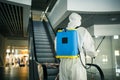 Portrait of a man in a sanitizing disifection suit holding spray near the escalator in an empty shopping mall. A volunteer