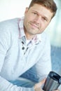 Portrait of man smiling and sitting on couch holding a flask. Attractive male inside his home with a travel coffee mug Royalty Free Stock Photo