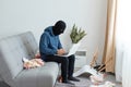 Portrait of man wearing blue hoodie and black robber mask, breaking flat, sitting on sofa and opening laptop, stealing modern