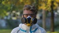 Portrait of man in white protective suit, glasses and respirator looking at camera on city street background. Virologist