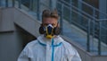 Portrait of man in white protective suit and respirator looking at camera on city street background. Virologist or