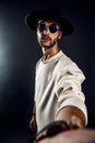 Portrait of a man in a wide-brimmed hat and sunglasses looking at camera while taking selfie on dark background. Royalty Free Stock Photo