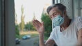 Portrait of masked senior woman in late 90s waving from home window. Elderly woman in protective mask looks out the