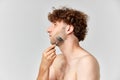 Portrait of matue man with curly hair, standign shirtless and doign face massage with roller against grey studio