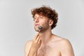 Portrait of matue man with curly hair, standign shirtless and doign face massage with roller against grey studio