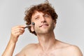 Portrait of matue man with curly hair, standign shirtless and doign face massage with roller against grey studio