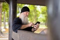 Mature handsome bearded hipster man using phone while sitting on wooden bench outdoors Royalty Free Stock Photo