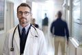 Portrait Of Mature Male Doctor Wearing White Coat With Stethoscope In Busy Hospital Corridor Royalty Free Stock Photo
