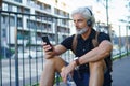 Portrait of mature man with headphones sitting outdoors in city, using smartphone. Royalty Free Stock Photo