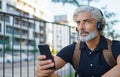 Portrait of mature man with headphones sitting outdoors in city, using smartphone. Royalty Free Stock Photo