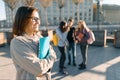 Portrait of mature smiling female teacher in glasses with clipboard, outdor with a group of teenagers students, golden hour