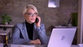 Portrait of middle-aged blonde short-haired businesswoman in glasses gladly talking on cellphone in office.