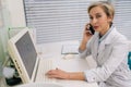 Portrait of middle-aged Caucasian female doctor in white coat talking on smartphone in hospital clinic sitting at desk Royalty Free Stock Photo