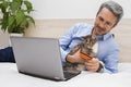 The handsome successful successful man with domestic cat lying on the sofa bed indoor and on-line shopping on a laptop. Royalty Free Stock Photo