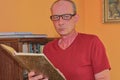 Middle aged man is reading book in living room. Mature man is standing next to bookcase.